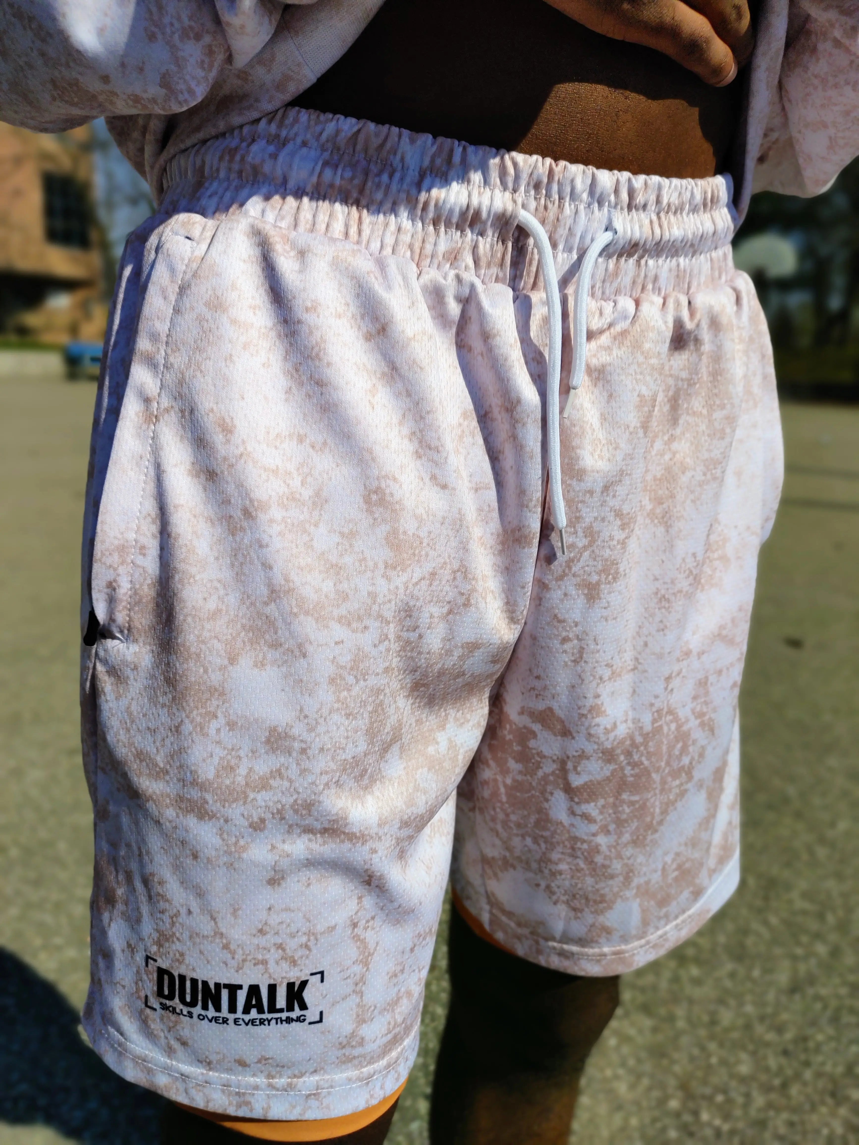 White basketball shorts with pink mud design, pockets, orange compression shorts underneath and tightening string at waist. Words on shorts say 
