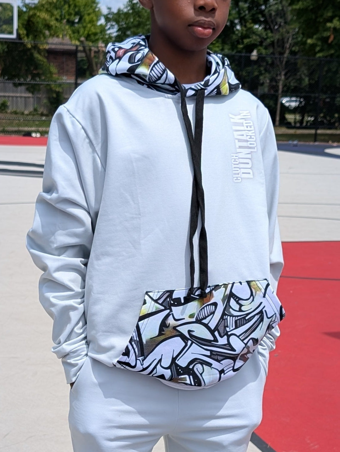 Person wearing a white hoodie and pants on a basketball court with trees in the background