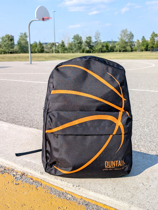 Front view of Black backpack with the seems of a basketball as the design. The bag is on a concrete surface with a basketball hoop in the background.
