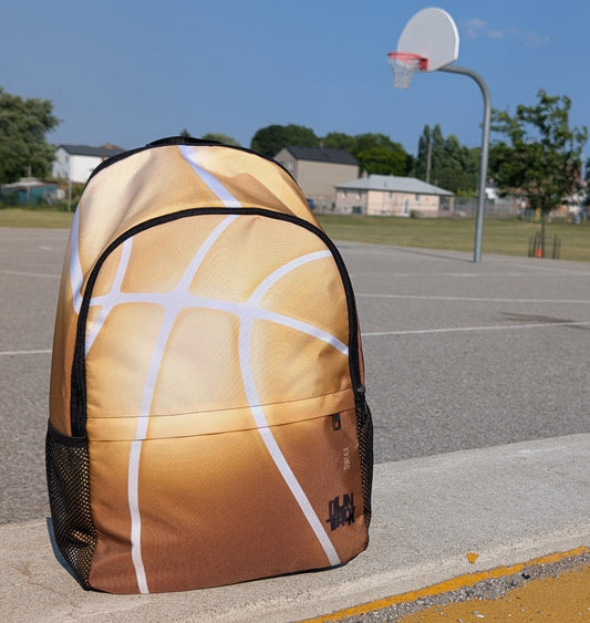 Backpack with the seems of a basketball as the design. Colours are a gradient of gold at the top to bronze at the bottom.  The bag is on a concrete surface with a basketball hoop in the background.