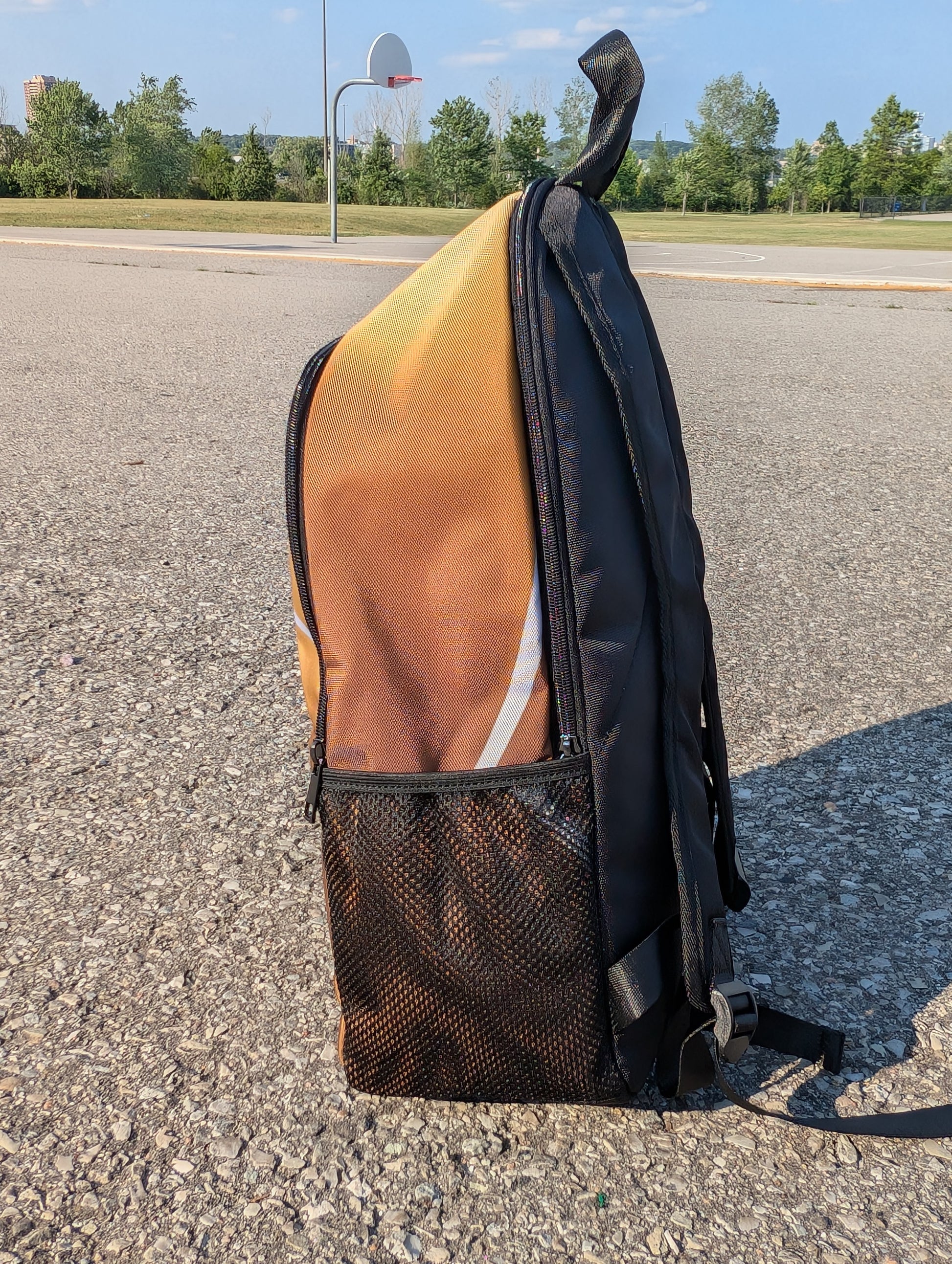 Side view of a backpack with the seems of a basketball as the design. Colours are a gradient of gold at the top to bronze at the bottom.  The bag is on a concrete surface with a basketball hoop in the background.
