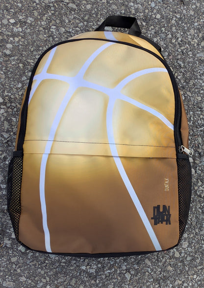 Backpack with the seems of a basketball as the design. Colours are a gradient of gold at the top to bronze at the bottom.  The bag is on a concrete surface.