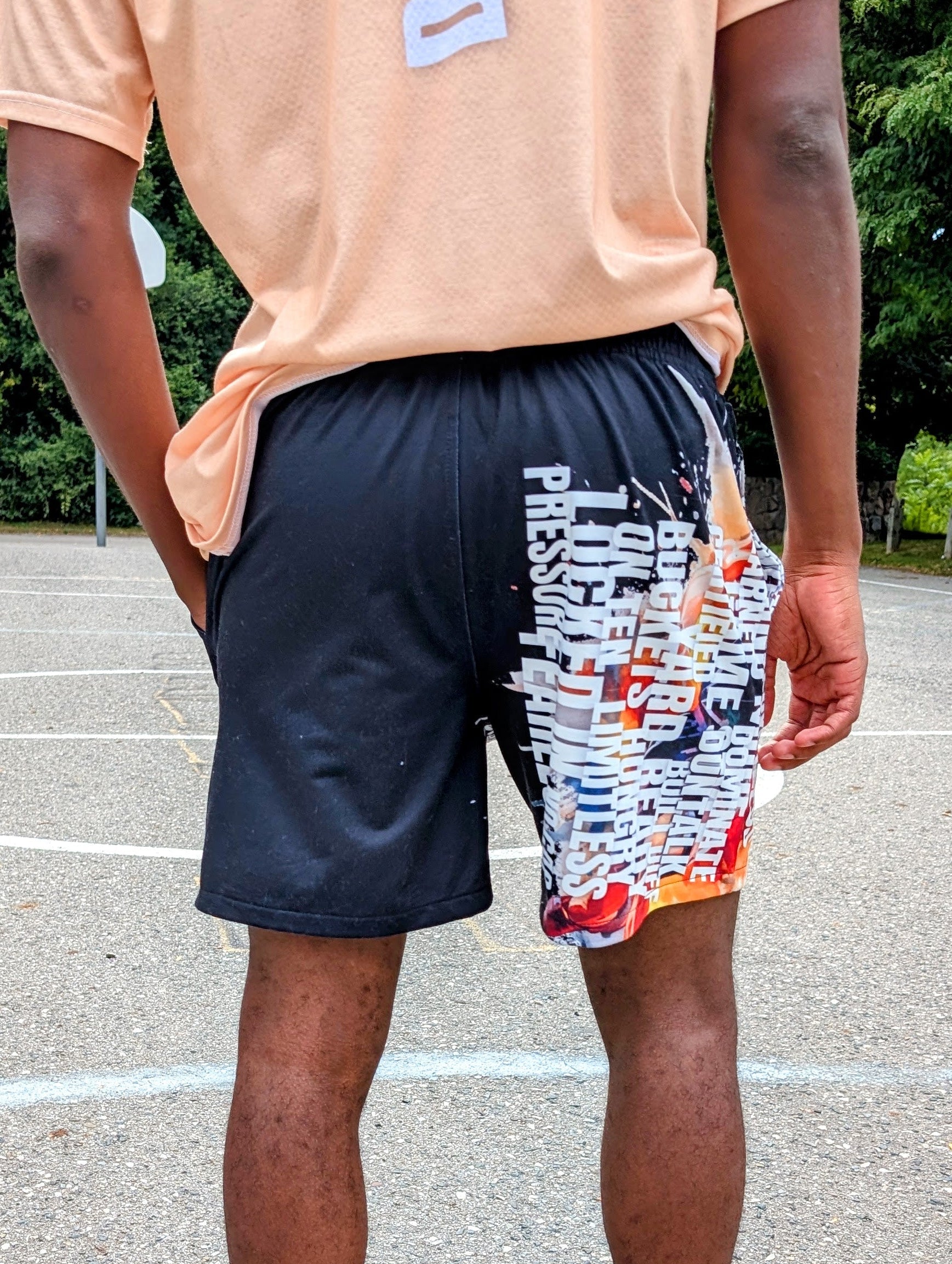 Person wearing a peach shirt, black shorts with colorful design, and black sneakers on a basketball court.